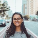 A young woman with glasses smiling on a city street, embracing urban lifestyle.